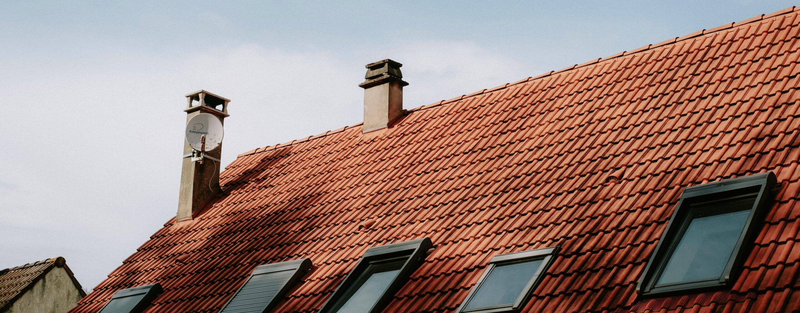 A picturesque red tiled roof featuring chimneys and skylight windows against a blue sky.